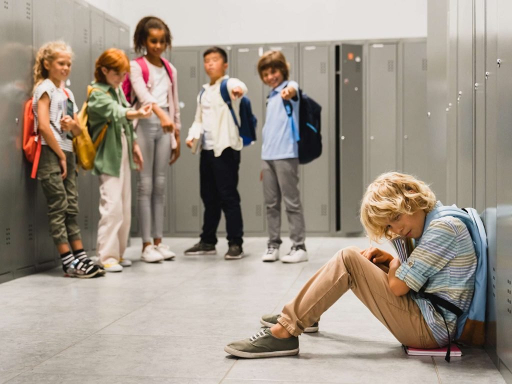 Sad crying schoolboy pupil sitting on the floor at the school hall while his classmates teenagers laughing at him, bullying and torturing him. Social exclusion problem
